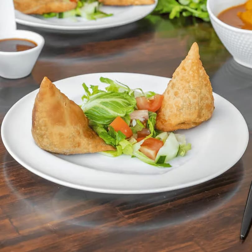 Two golden-brown samosas with a fresh side salad of lettuce, tomatoes, and cucumbers on a white plate.