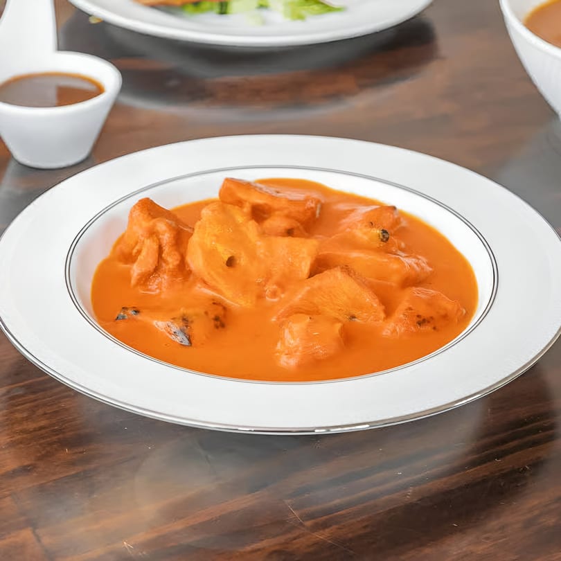 A white bowl of Indian paneer butter masala with a side of brown sauce.