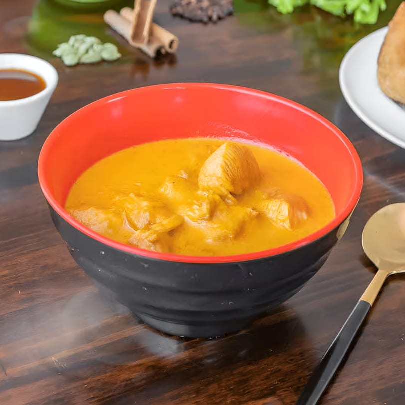 A vibrant orange Indian curry with chicken pieces in a black and red bowl, with a gold spoon.